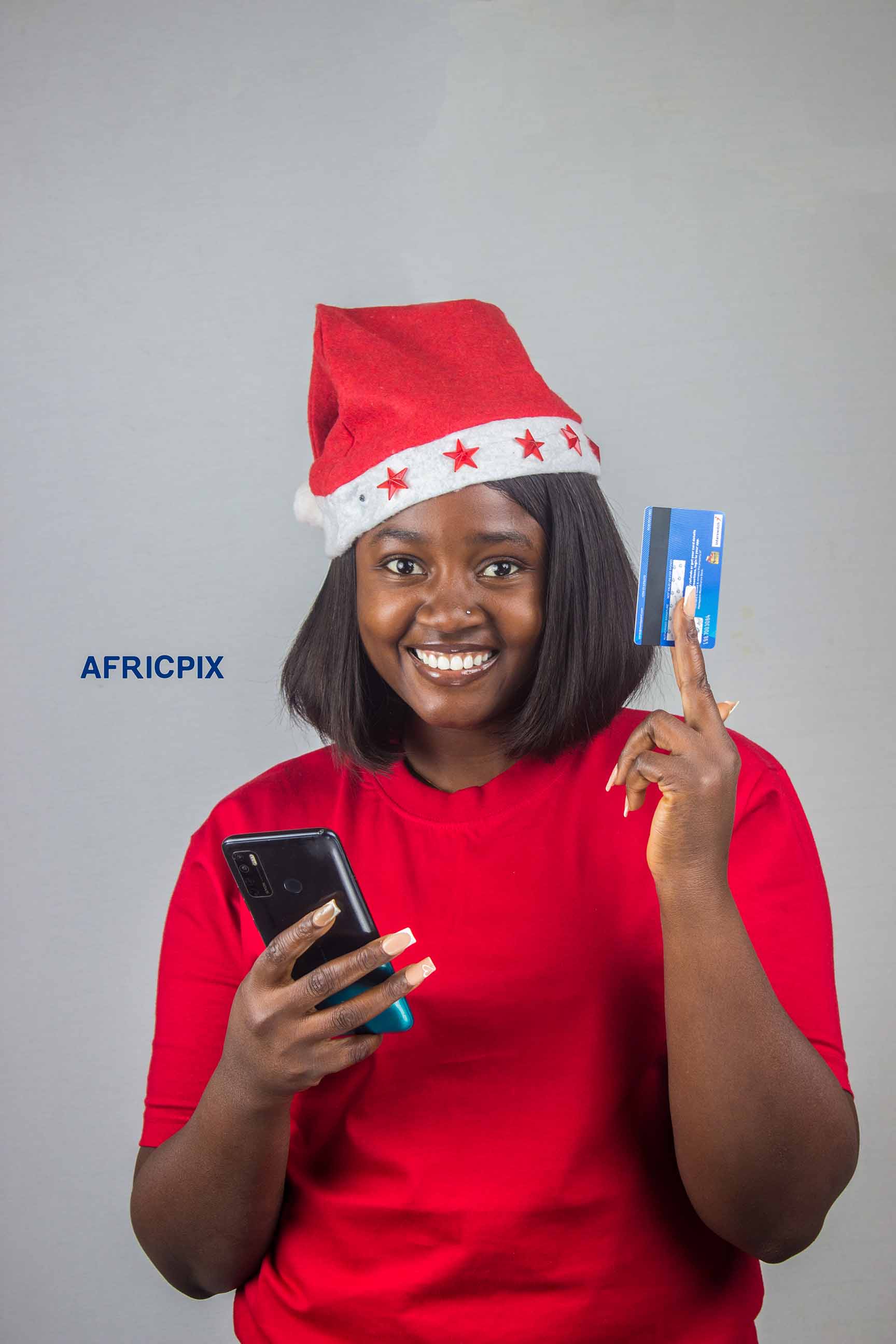 A happy Nigerian African woman wearing a Christmas cap, holding an ATM card and phone, smiling joyfully.